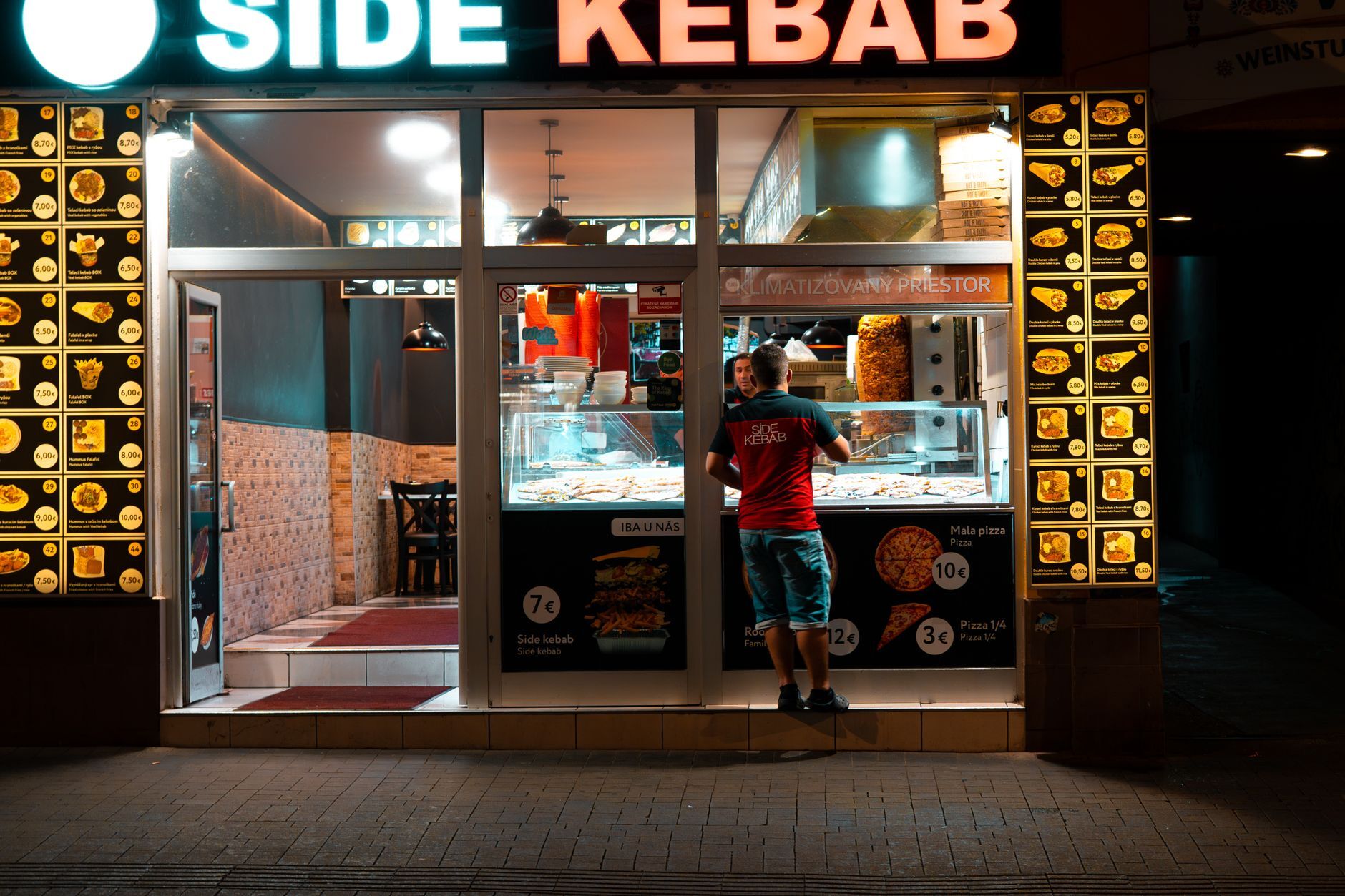 Illuminated kebab shop façade at night with menu displays and customer. Photo by Lukas Kosc on Pexels