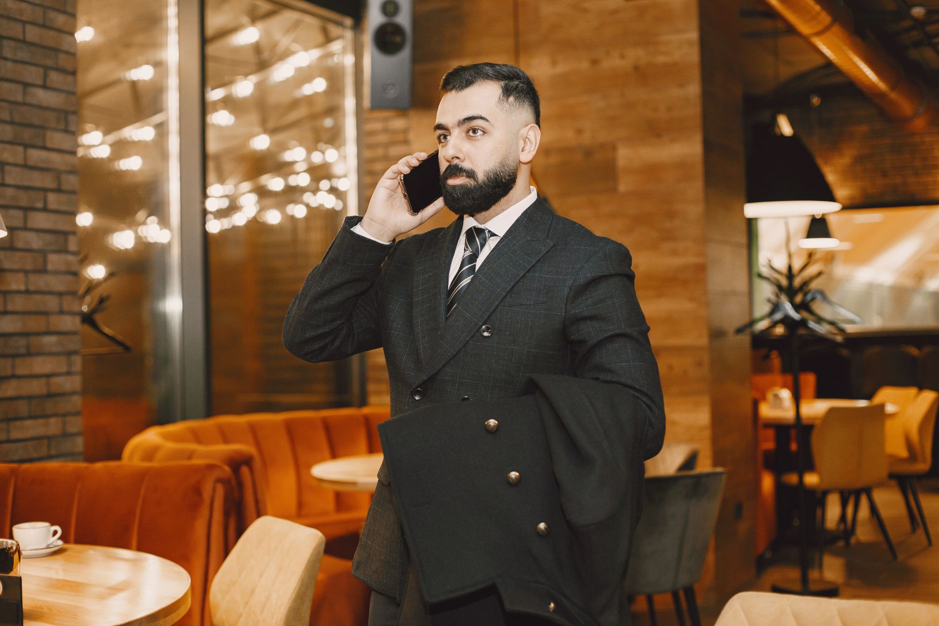 Bearded man in black suit talking on phone in a chic cafe setting. Photo by Gustavo Fring on Pexels