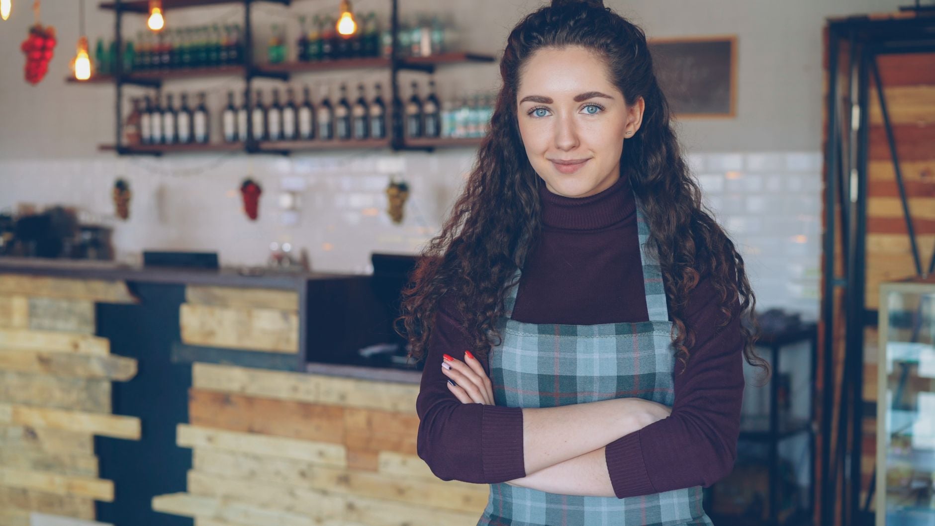 Confident café owner stands with arms crossed inside a rustic, cozy coffee shop. Photo by Vitaly Gariev on Pexels