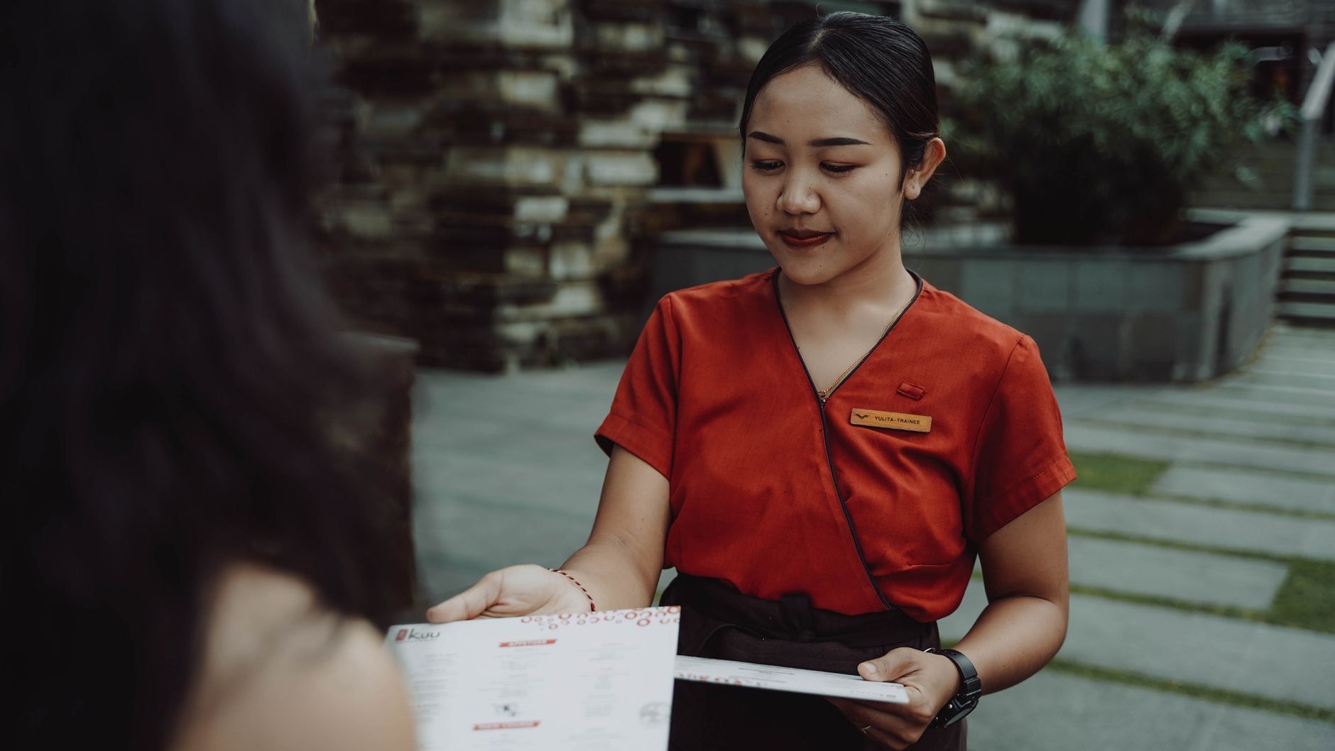 A waitress in a red uniform handing a menu to a guest at an outdoor restaurant. Photo by Bhefan Tedjo on Pexels